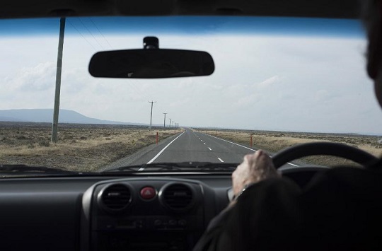 Image of a Long journey ahead concept with man driving a car in an over the shoulder perspective looking at the long road stretching into the distance through the windscreen
